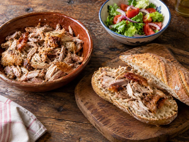 Pollo asado desmigado en un bol, con un bocadillo a medio montar y un cuenco de ensalada al lado